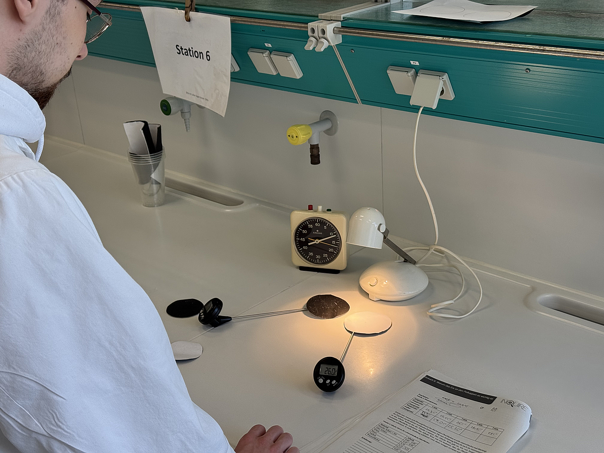 Schmelzende Polkappen und der Albedo-Effekt Ein Schüler in Kittel steht an einer Laborbank und betrachtet einen Versuch zum Albedo-Effekt, bei dem eine eingeschaltete Lampe auf eine weiße sowie eine schwarze Fläche leuchtet und jeweils die Temperatur gemessen wird.