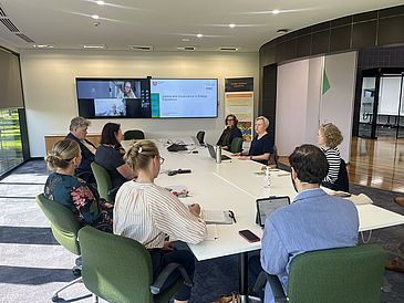 In a conference room, people are sitting around a table listening to Stefanie Baasch’s presentation; other online participants can be seen on screens on the wall.