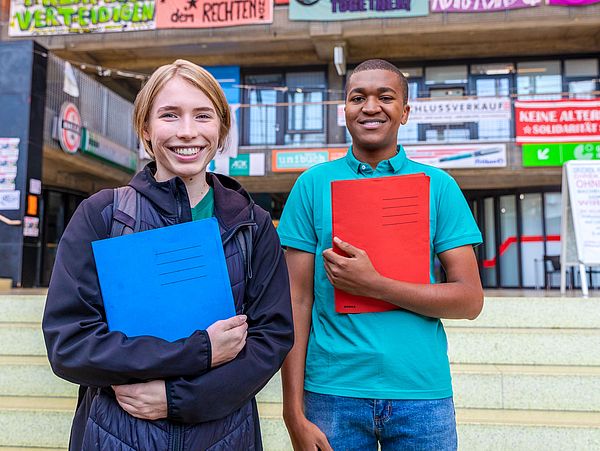 Zwei Studierende mit Mappen in der Hand lächeln in die Kamera