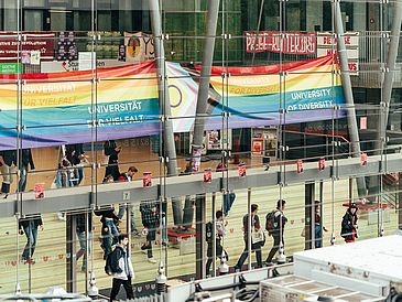 Menschen in der Glashalle, Pride-Flagge im Hintergrund.