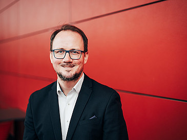 Physicist Andreas Sentef stands in front of a red wall in a dark suit and smiles.