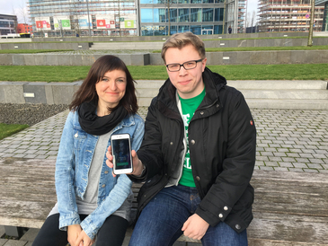 Junge Frau und junger Mann sitzen auf einer Treppenstufe und halten ein Smartphone in die Kamera
