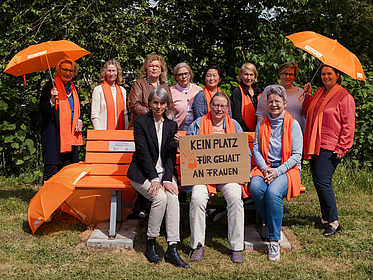 Eine Gruppe von Frauen hat sich um eine orangefarbene Bank versammelt. Eine Frau hält ein Schild mit der Aufschrift "Gegen Gewalt an Frauen".