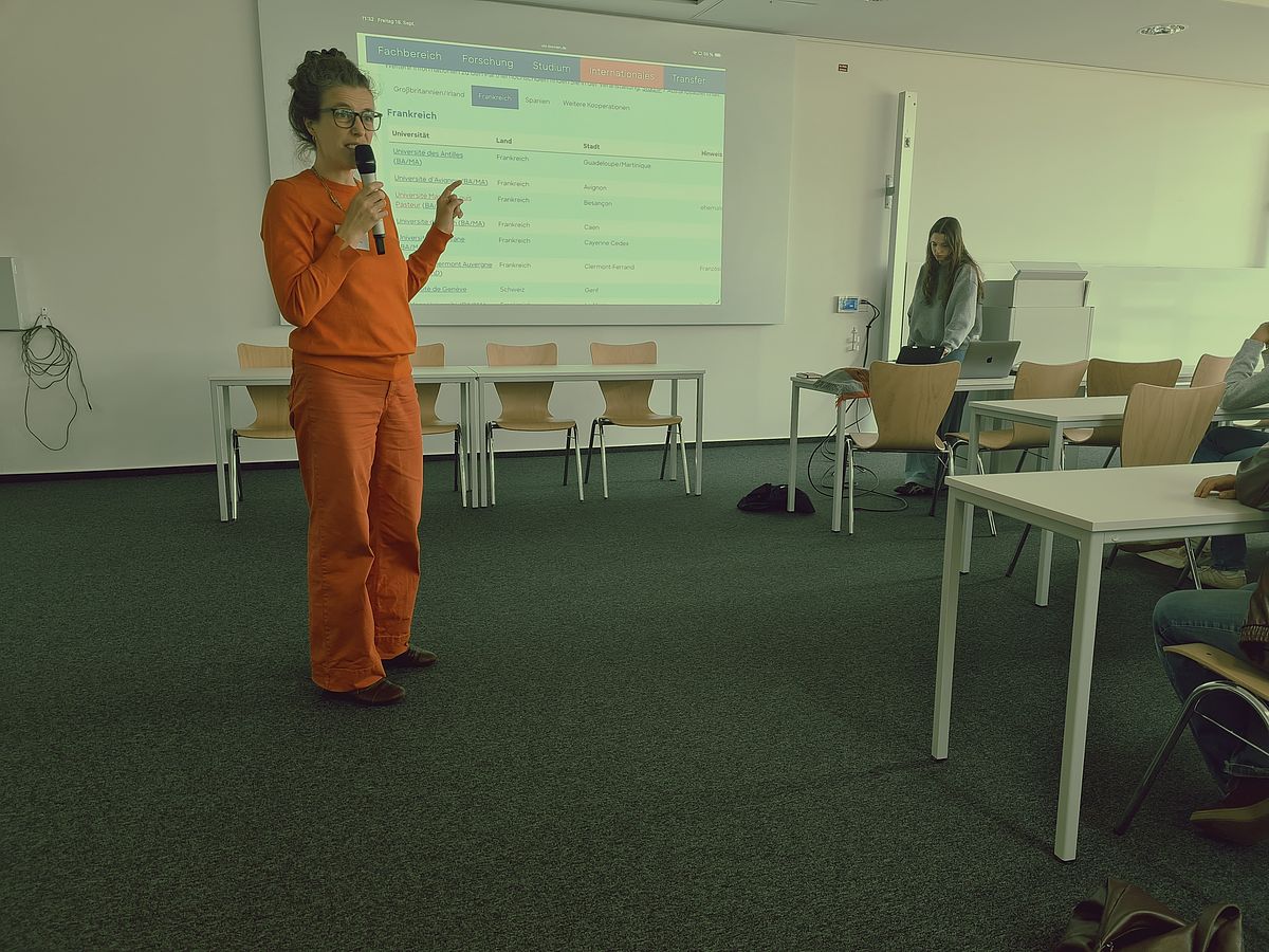 Im Vordergrund steht eine referendierende Person; im Hintergrund wird eine Präsentation vorbereitet.