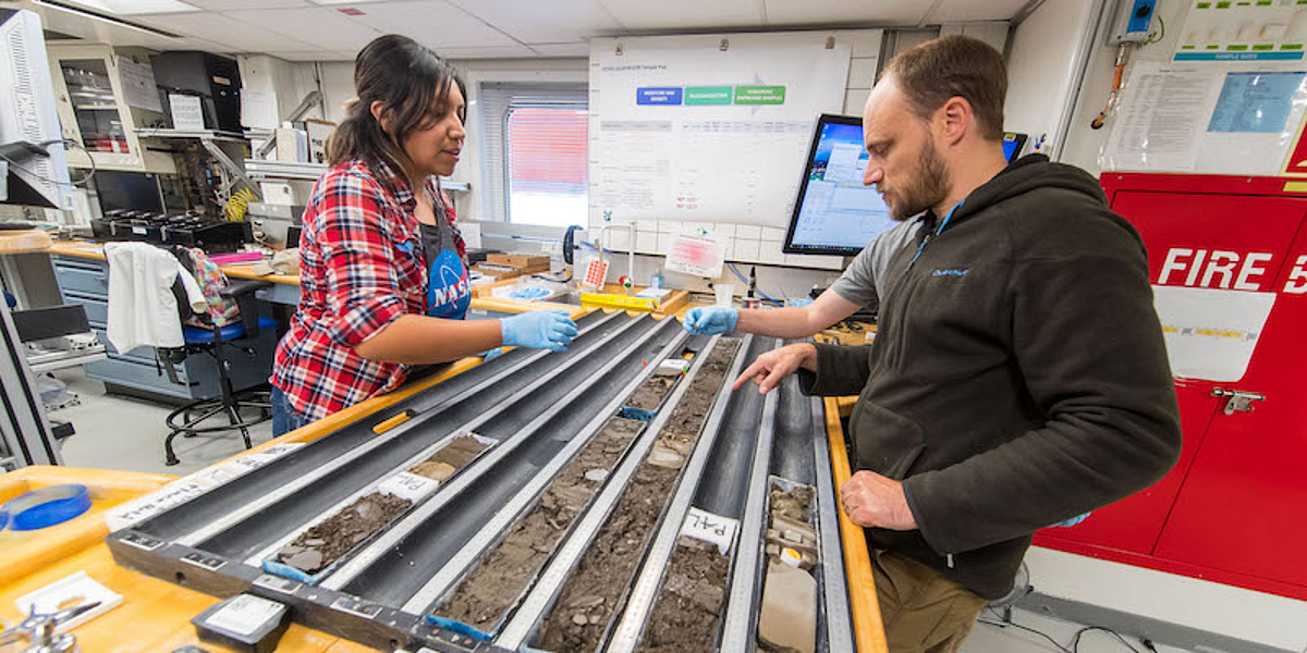 A female scientist and two male scientists stand in the laboratory of the drill ship Joides Resolution and sample rock cores in sawn open plastic tubes lying on the laboratory table.