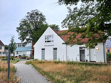 Ein gepflasterter Fußweg führt durch eine wilde Wiese zu einem weißen Haus, auf dem "Fahrradverleih" steht