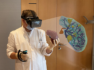 Man in white coat, mask and VR glasses looks at 3D-printed human liver he holds in his left hand.