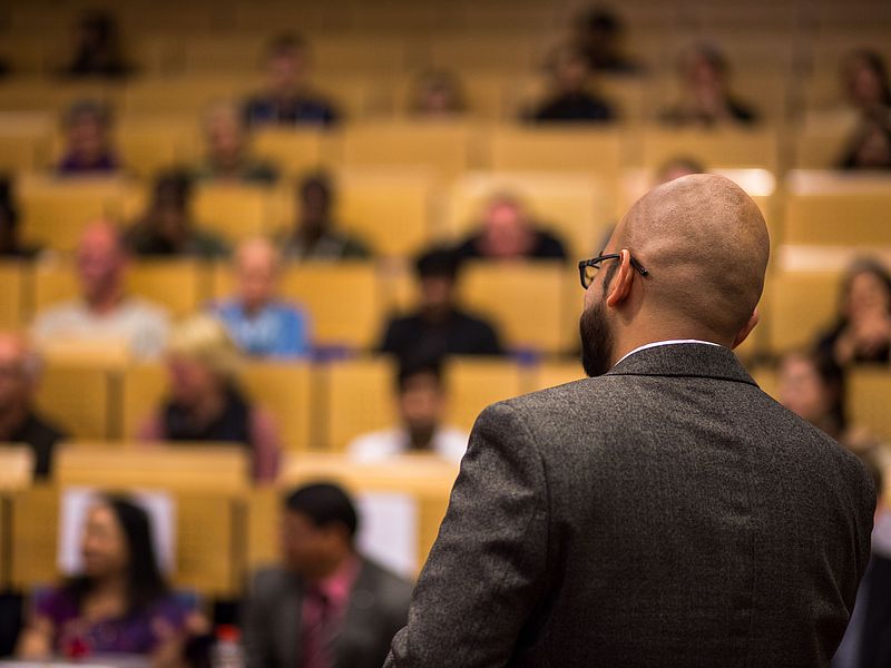 A lecturer speaks to an audience in a packed lecture hall.