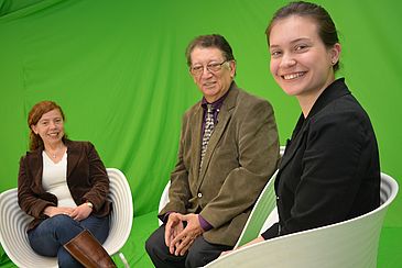 Drei Menschen sitzen in weißen Stühlen vor grünem Hintergrund