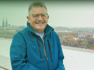Ein Foto von Jeremy Hookway. Er sitz auf einer Dachterasse, hinter ihm die Stadt Bremen. Er lächelt in die Kamera