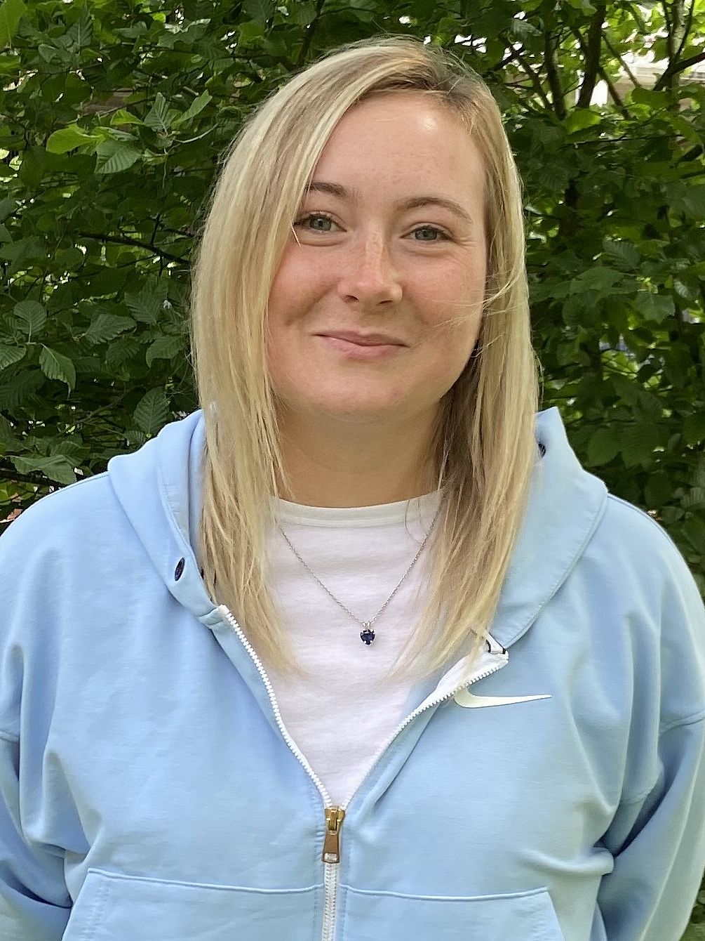 Lena ist eine junge Frau mit blonden Haaren. Sie steht vor einem grünen Busch und lächelt freundlich in die Kamera.