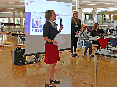 Frau sprich in das Mikro vor einer Tafel in der Mensa Uni Bremen