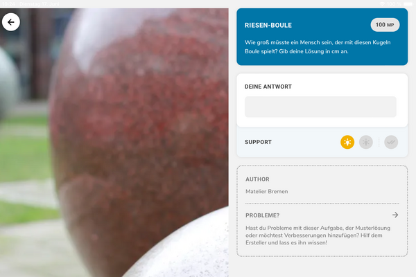 Screenshot aus der App MathCityMap. Links ist ein Bild, das zwei Marmorkugeln angeschnitten zeigt, die eine davon unscharf. Rechts ist eine Aufgabenstellung zu lesen: "Riesen Boule - Wie groß müsste ein Mensch sein, der mit diesen Kugeln Boule spielt? Gib deine Lösung in cm an.