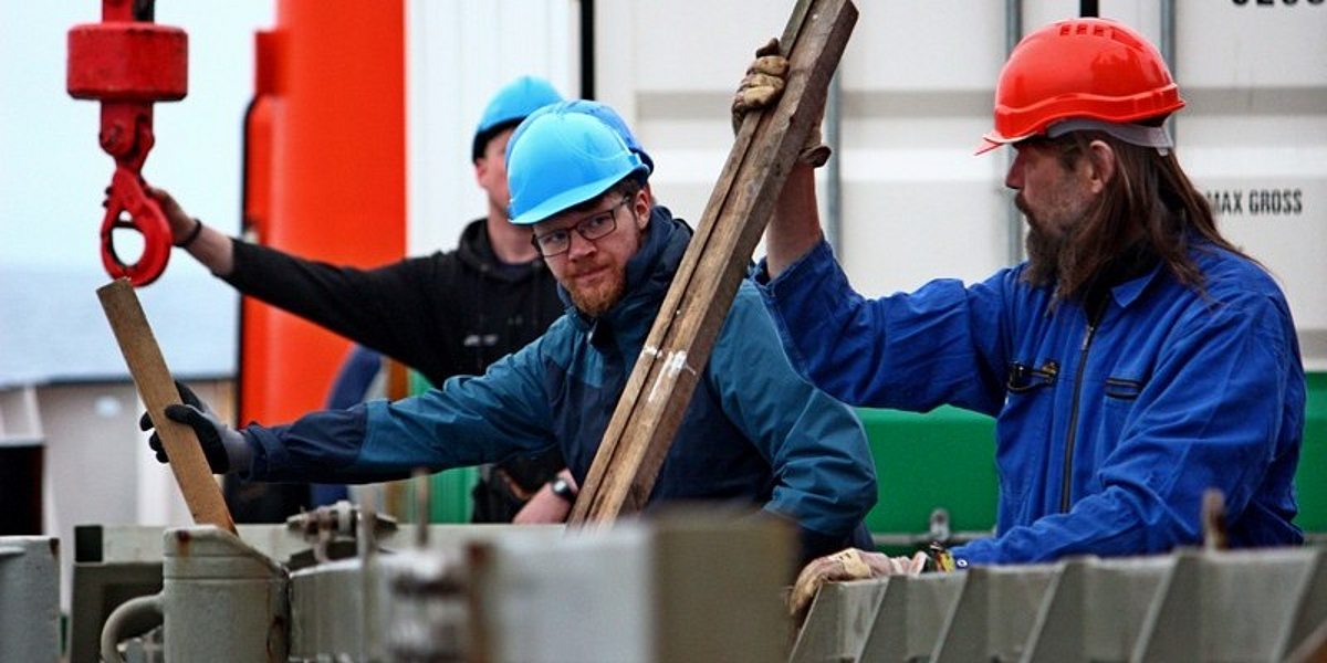 Three men in blue work clothes with hard hats and protective gloves stand on the deck of the research vessel Meteor. They hold wooden slats in their hands and are waiting for the arrival of the gravity corer.