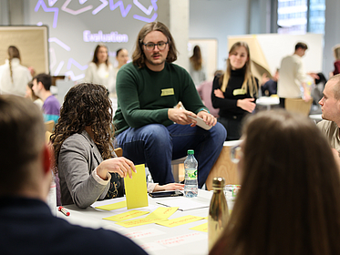 Mehrere Studenten um einen Tisch eine Studentin hält eine gelben Klebezettel.