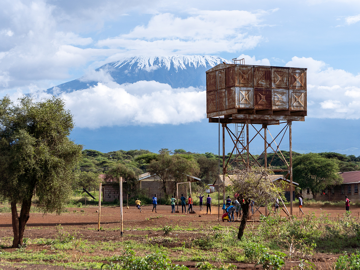 In the foreground, a large rusty container stands on an approximately eight-meter-high steel frame. In front of it, teenagers are playing soccer. Behind them, Mount Kilimanjaro in Tanzania is visible, seen from the Kenyan side.