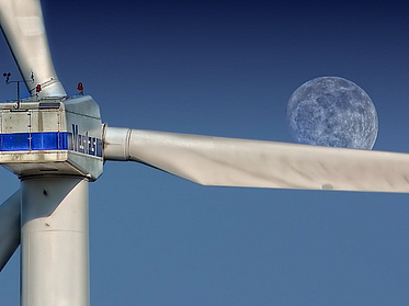 Close-up of the pulpit of a wind turbine, behind one of the blades the moon in the blue sky