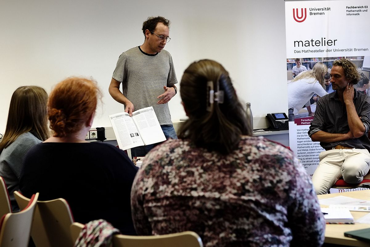 Ein Mann steht vor einer Gruppe von sitzenden Menschen und scheint etwas zu sagen. Er hält dabei ein aufgeschlagenes Heft in der Hand. Im Hintergrund ist ein Aufsteller mit der Überschrift "matelier" zu sehen.