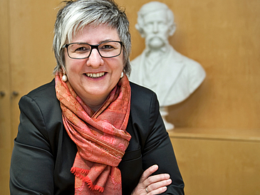 Portrait of a woman with short gray hair and glasses. She wears a black jacket with a red scarf.