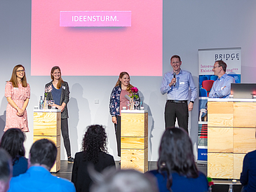 Five people stand at three standing desks in front of a screen that reads "Ideensturm" [Ideas Storm]. One person holds a microphone in their hand.