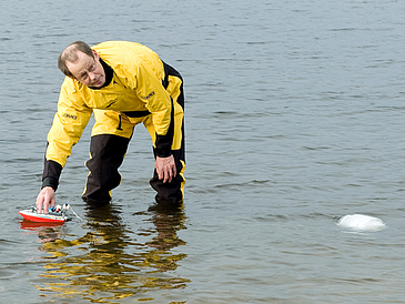 Mann in Trockenanzug steht knietief im Wasser während ein Spielzeugboot versucht, einen Eisblock zu ziehen.