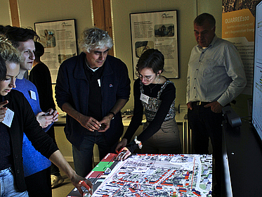 Workshop-Teilnehmer:innen stehen um einen Tisch, auf den die Grundrisse einer Stadt projiziert sind