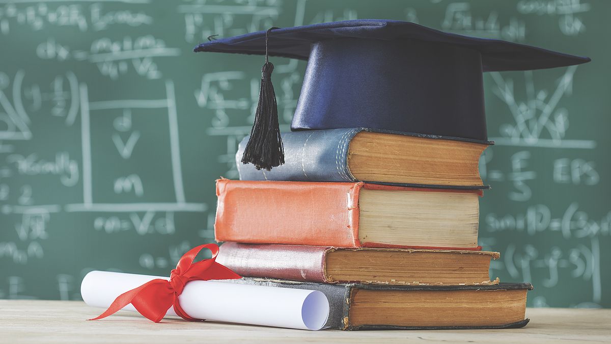 Stack books graduate hat and diploma in front of green chalkboard Duale Promotion
