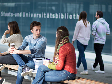 Studierende sitzen vor dem Eingang der Bibliothek.