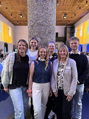Sechs Personen stehen als Gruppe in einem Saal vor einer Steinsäule.