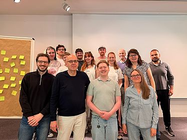 Eine Gruppe aus Leitenden und Teilnehmenden des Workshops "Metaphors Open" steht vor einer Tafel und einer Pinnwand.