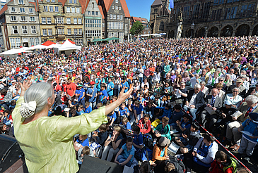 Dirigentin mit ausgebreiteten Armen und tausende Sänger auf dem Bremer Marktplatz