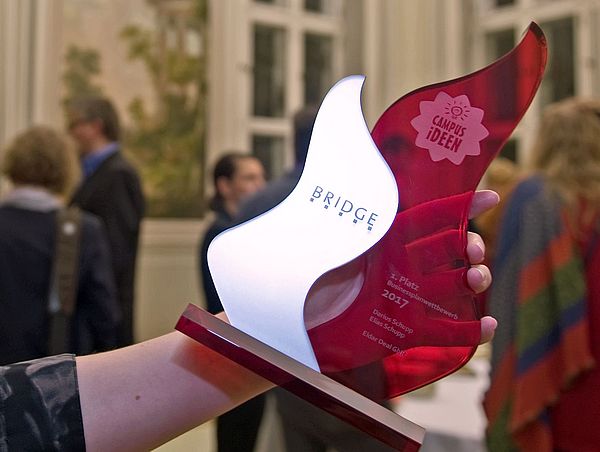 eine Hand hält einen Preis, der auf dem CAMPUS-Ideenwettbewerb von BRIDGE verliehen wird
