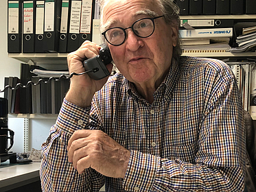 An elderly man sits at a desk and holds a phone with a cord to his ear.