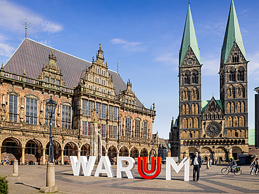 Das Wort WARUM steht in großen Buchstaben mit dem Uni-Logo stehen auf einem Marktplatz mit Menschen. Im Hintergrund Rathaus und Dom.