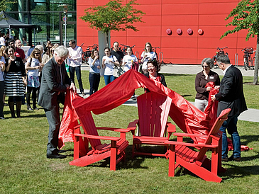 Vier Personen, zwei Männer und zwei Frauen, ziehen ein rotes Tuch von Stühlen ab 