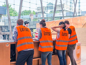 Vier junge Menschen stehen mit orangenen Warnwesten an einem Geländer und schauen hinab in eine große Halle. Auf ihren Westen steht das Wort "Lotse". Das Bild ist von hinten aufgenommen.