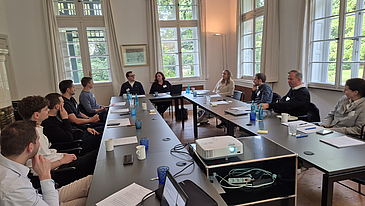 People are seated at tables arranged in a U-shape and are looking at Professor Klein.