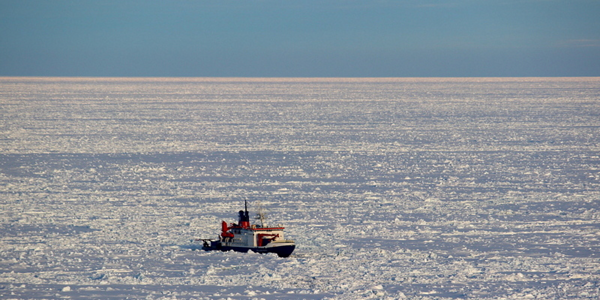 Aerial view of the research vessel Polarstern surrounded by sea ice. The sun is shining in the cloudless sky.