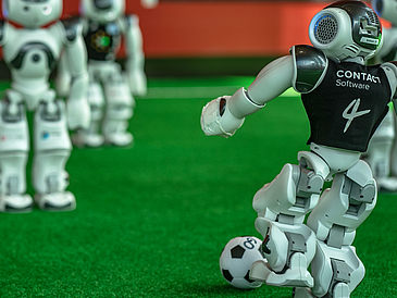 Roboter auf einem Spielfeld. Der rechte kickt den Ball.