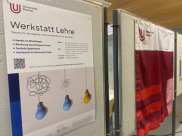 Links auf dem Bild hängt das Plakat der Werkstattlehre an einer Stellwand, rechts daneben eine Fahne der Universität Bremen im Corporate Design.