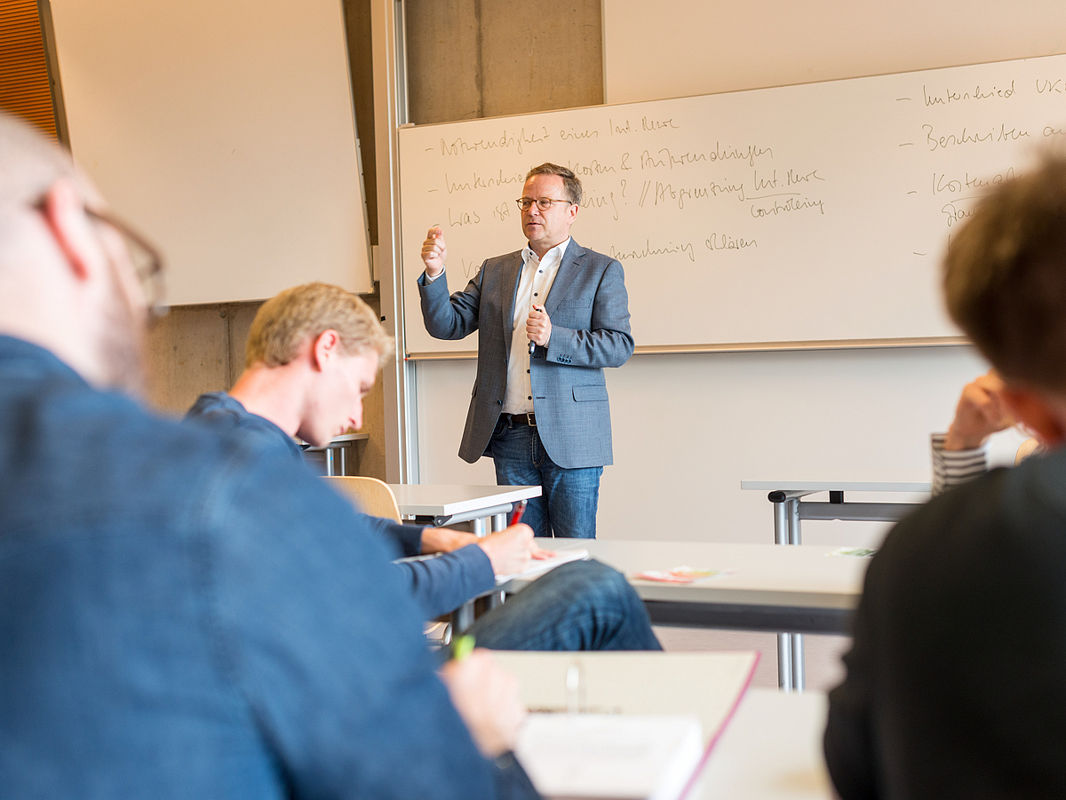 Ein Lehrender spricht vor Zuhörern in einem Seminarraum.