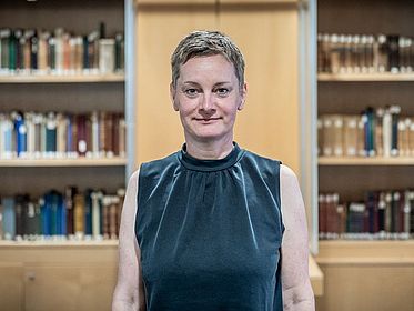 Dr. Barbara Aehnlich steht im Vordergrund des Bildes, dahinter sind Bücherregale zu erkennen.