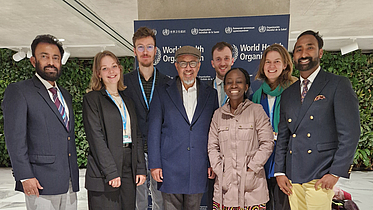 Matthew Willis und Anil Fastenau zusammen mit Kolleg:innen und dem WHO Director General Dr. Tedros Adhanom Ghebreyesus stehen vor einer Wand mit WHO Logo
