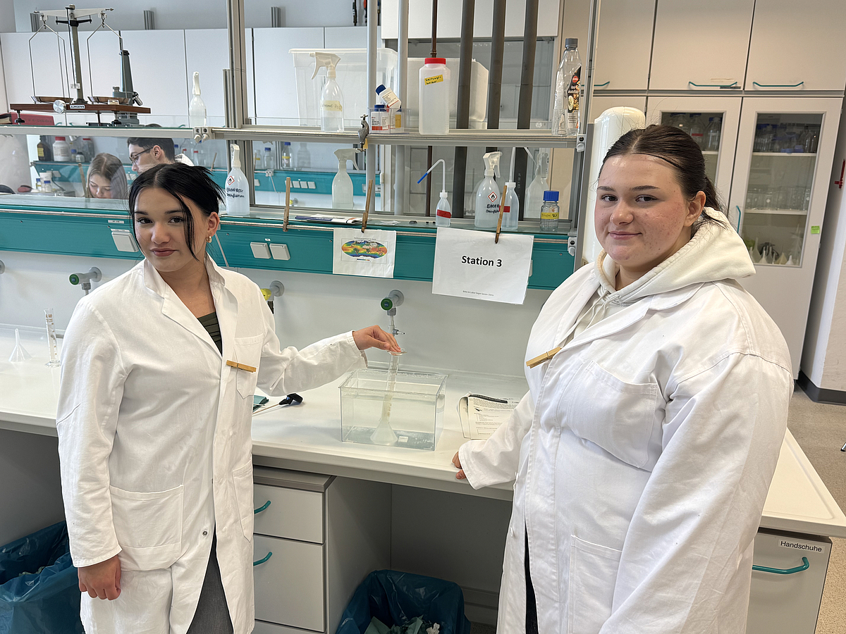 Ein Planspiel mit praktischen Elementen im Labor Zwei Schülerinnen in Kitteln stehen an einer Laborbank und schauen in die Kamera.