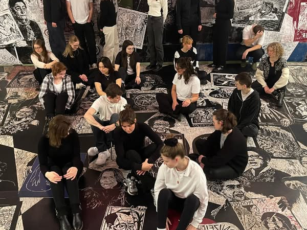 Eine Gruppe, warhscheinlich die Studierenden des Kunstrausches sitzen auf einem Boden, der übersäht ist mit schwarzweiß Zeichnungen, die jedoch als Teil des Bodens wirken