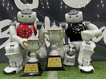 Zwei B-Human Roboter posieren glücklich mit ihren zwei neuen Pokalen.