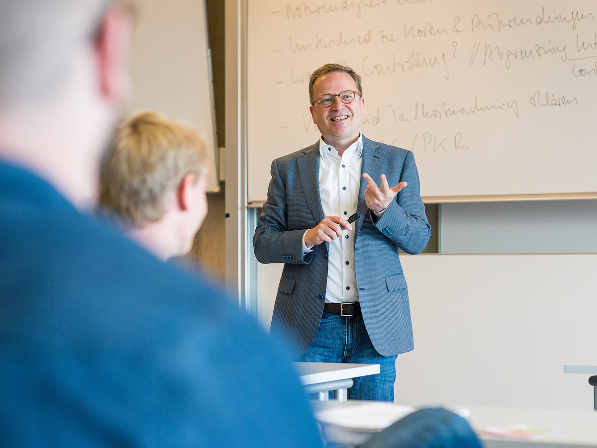 A teacher speaks in front of an audience in a seminar room.