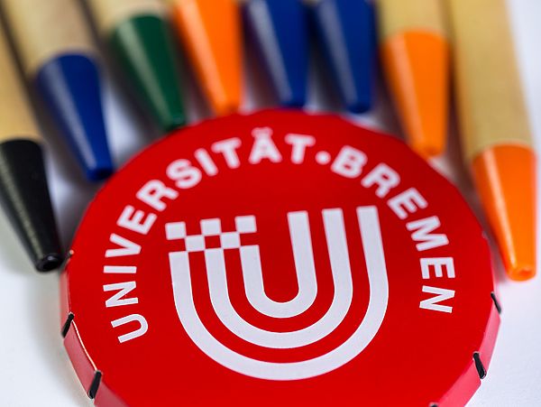 Eine Dose mit Universitätslogo liegt neben Stiften.