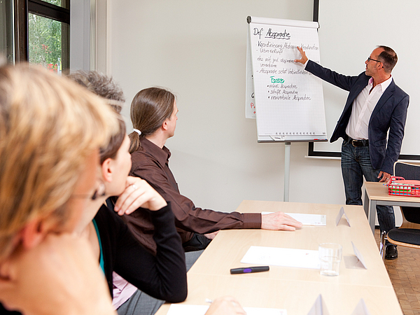 A teacher stands in front of an audience at a flipchart.