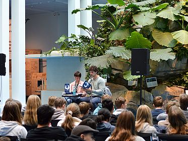 The picture shows a school reading of the work ‘Auf dem Tigerpfad’ (On the Tiger Trail) with author Jakob Graf and presenter Myriam Macé inside the Übersee Museum. In the foreground, many pupils are sitting and listening to the presentation.